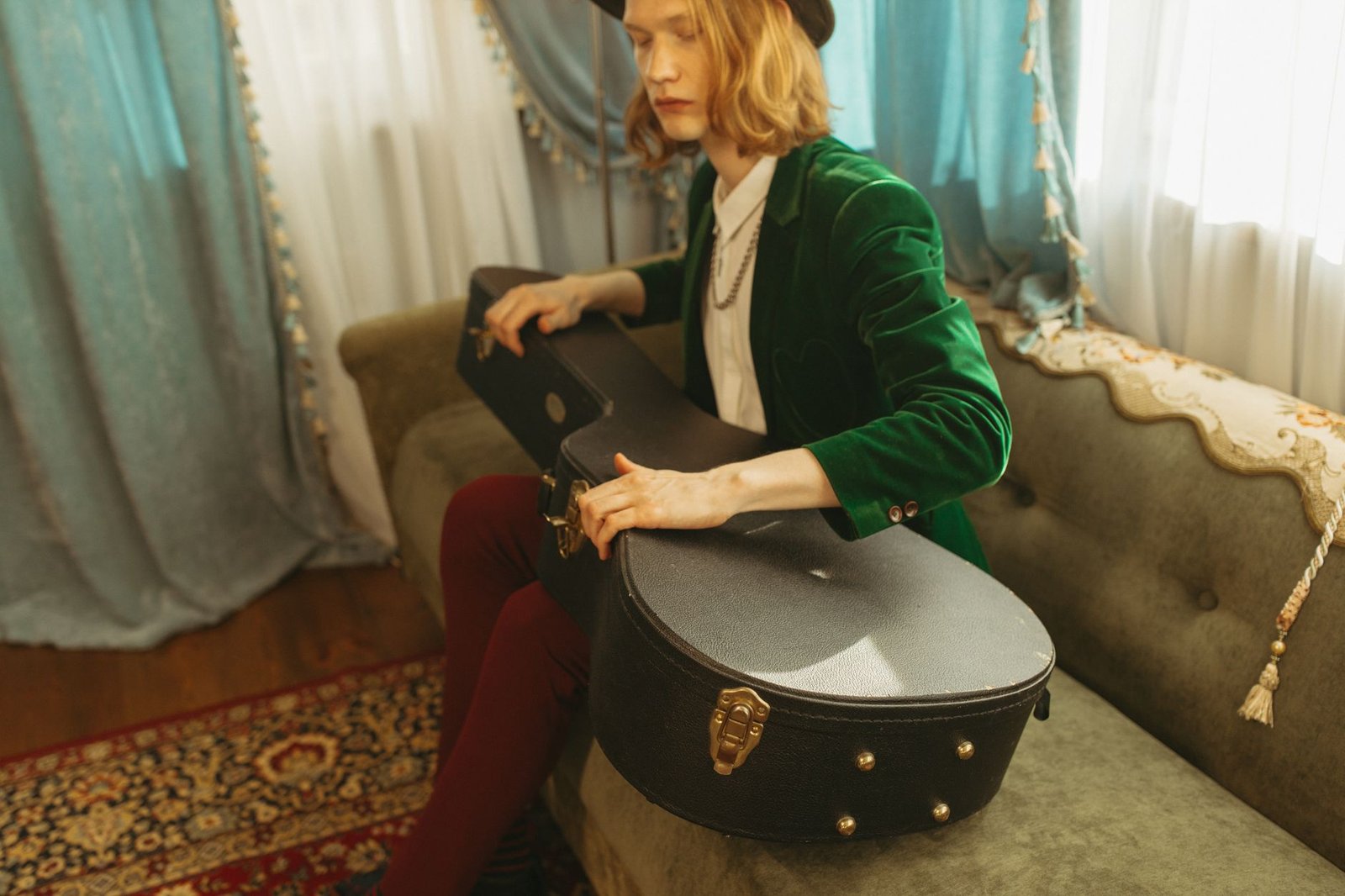 A person in a green velvet blazer sits on a sofa holding a guitar case indoors.