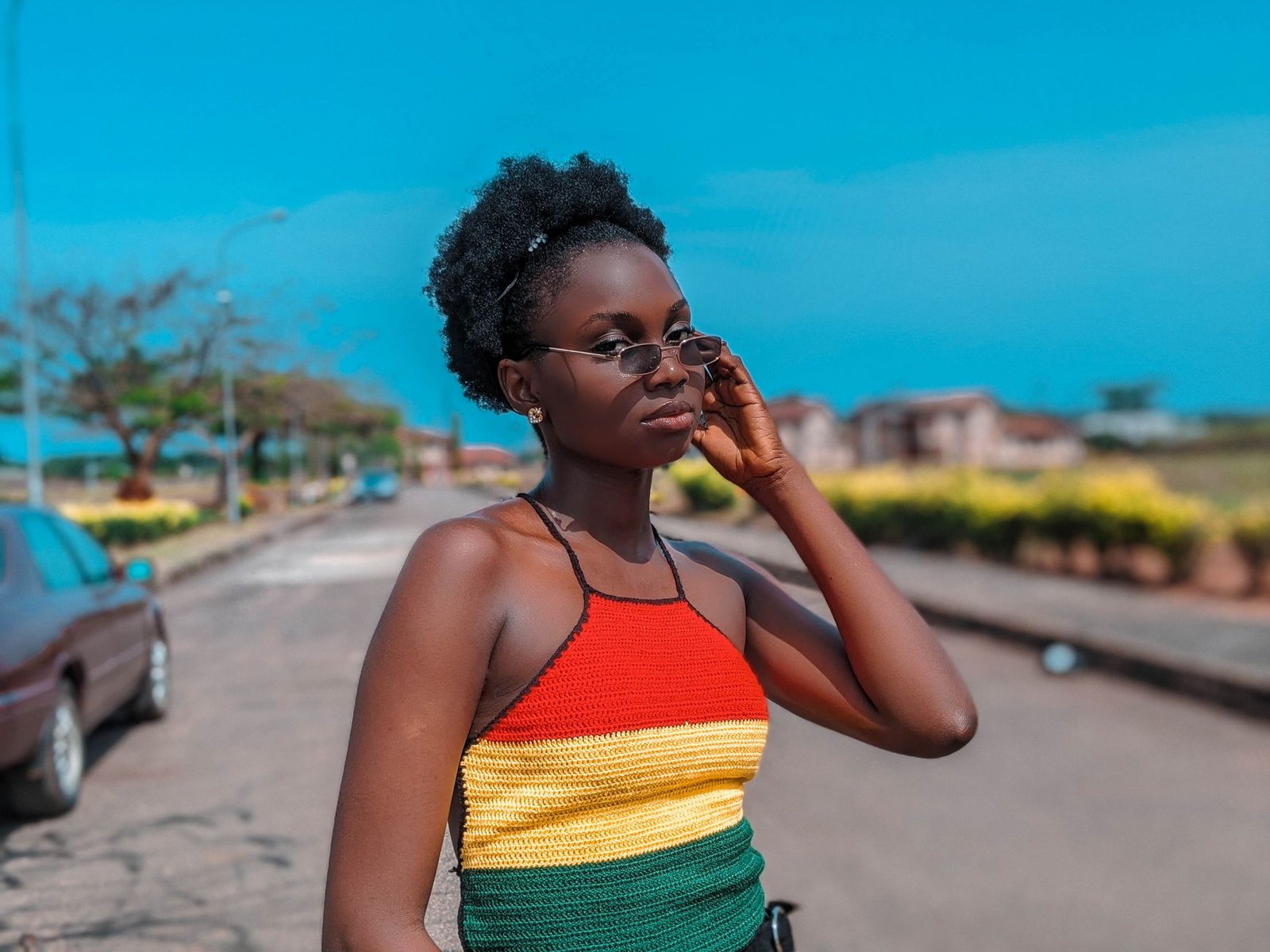 Confident young woman wearing sunglasses and colorful outfit poses stylishly on a sunny street.