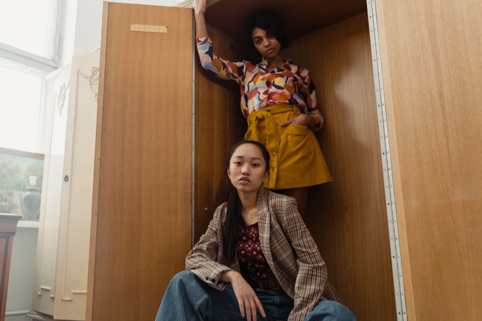 Two women in vibrant outfits posing inside a wooden cabinet, showcasing modern fashion.
