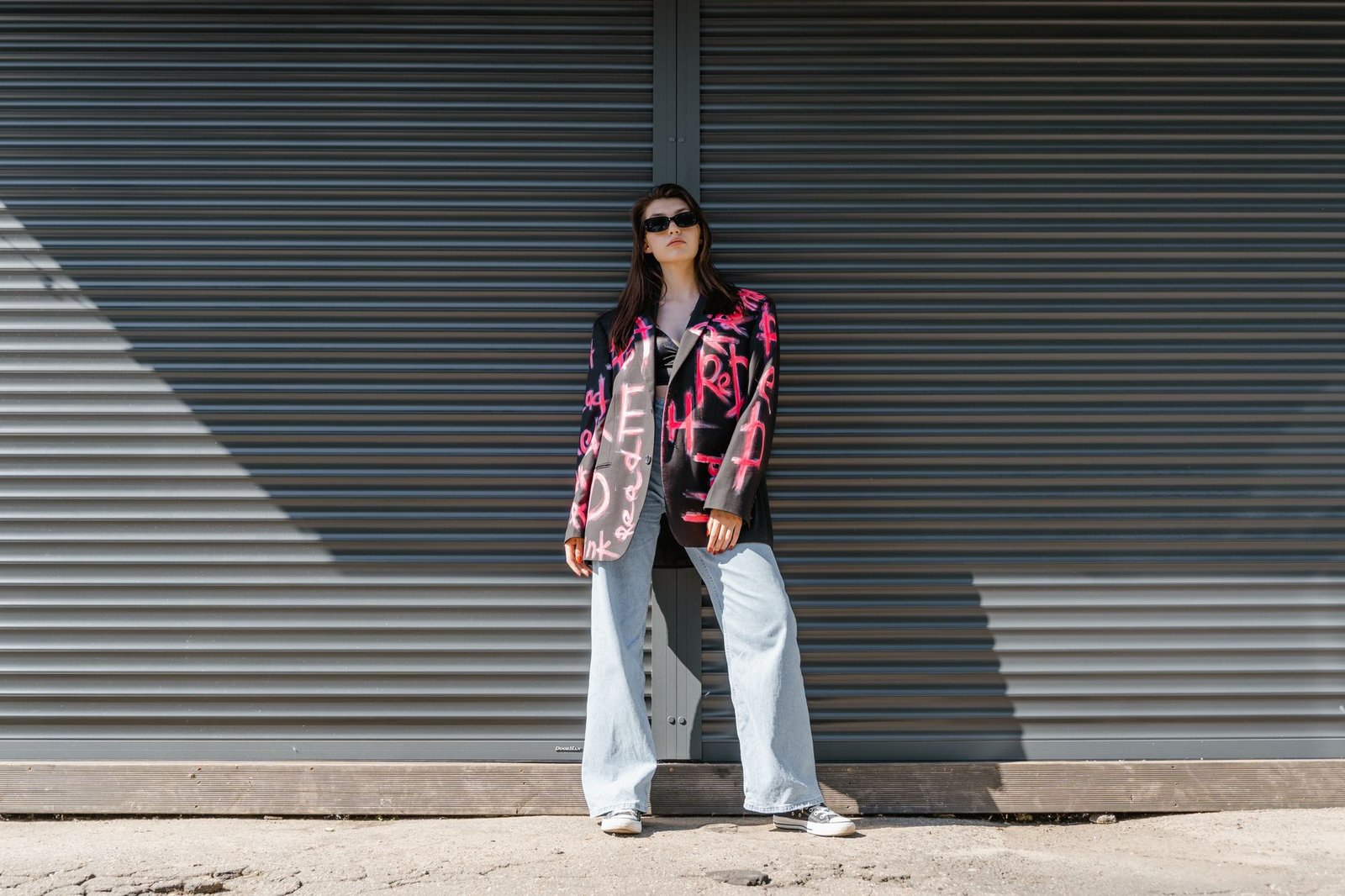 Stylish woman in a graffiti blazer and denim pants posing against a metal shutter.