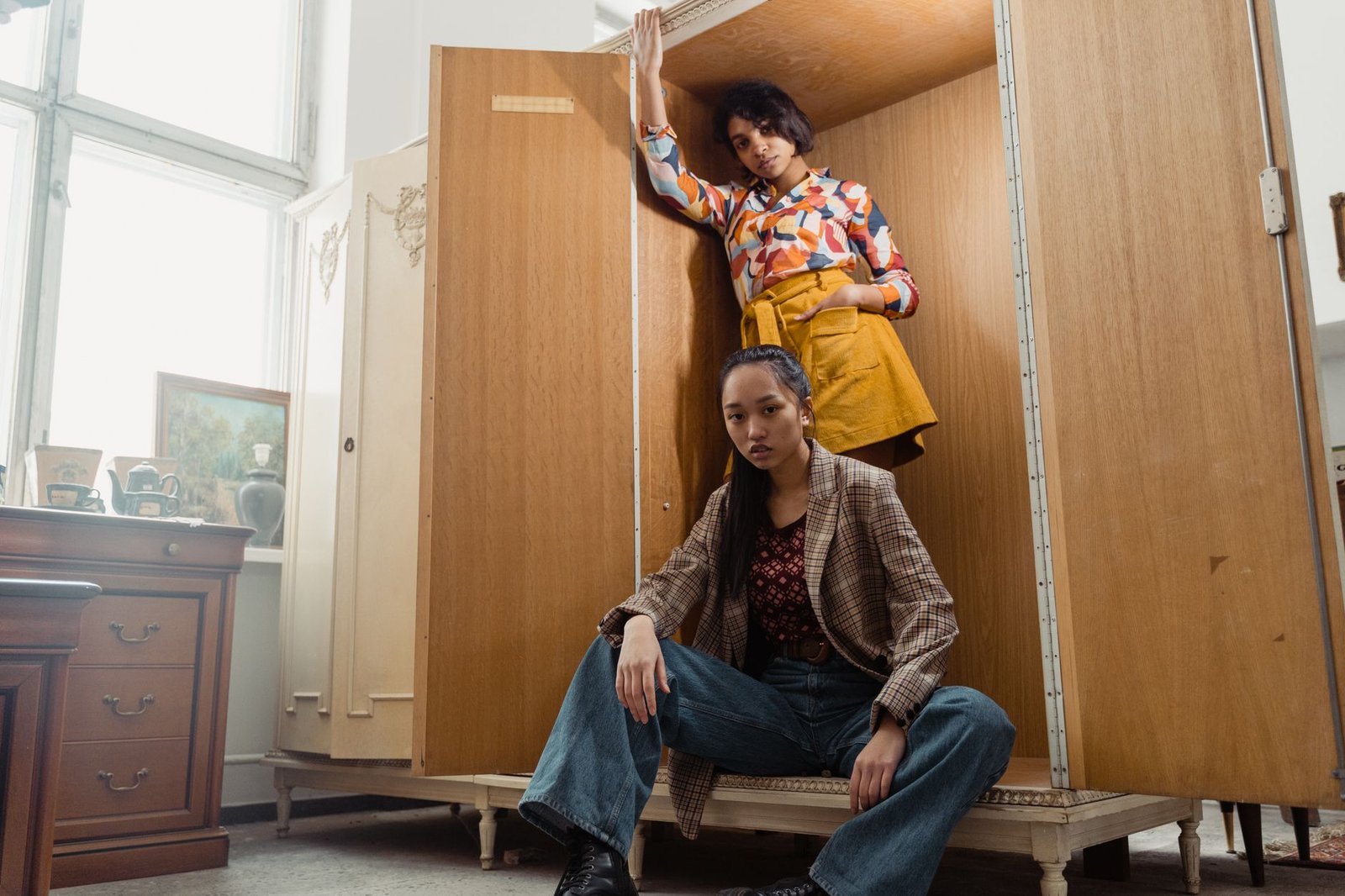 Two fashionable women posing creatively with a vintage wooden cabinet indoors.