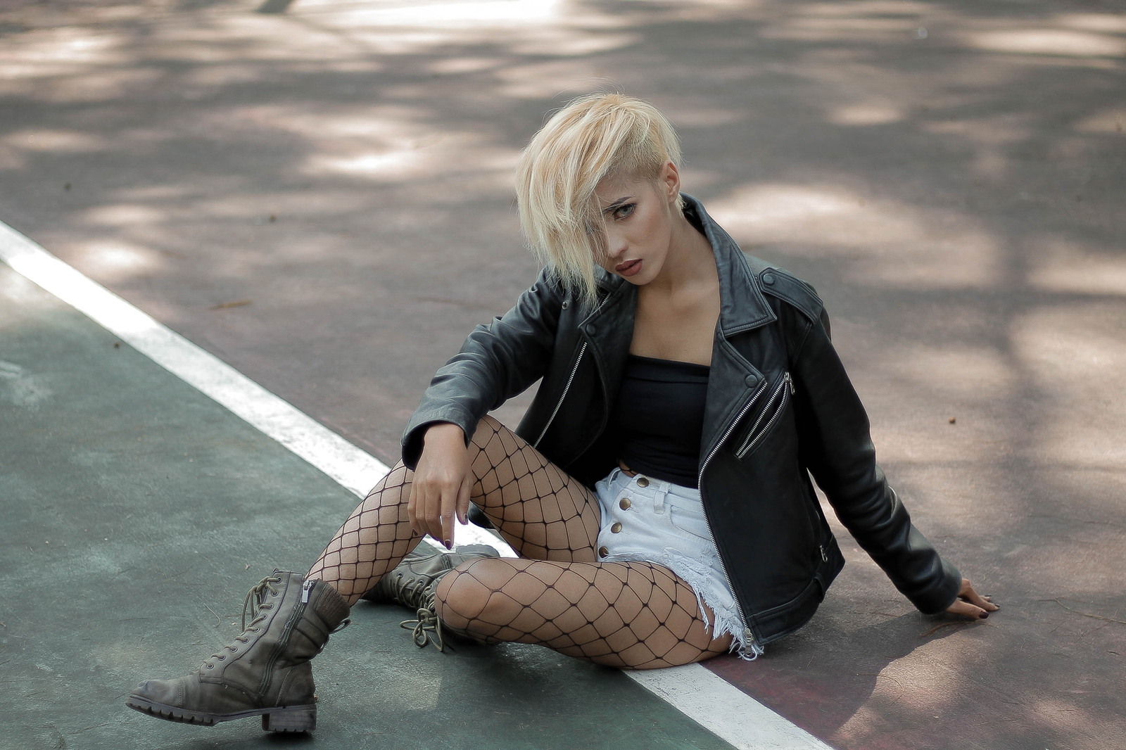 Fashionable woman in leather jacket and fishnet stockings posing on outdoor pavement.