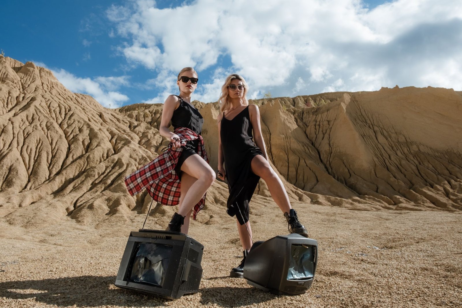 Two women in fashionable attire stand in a desert landscape with vintage televisions, striking a bold pose.