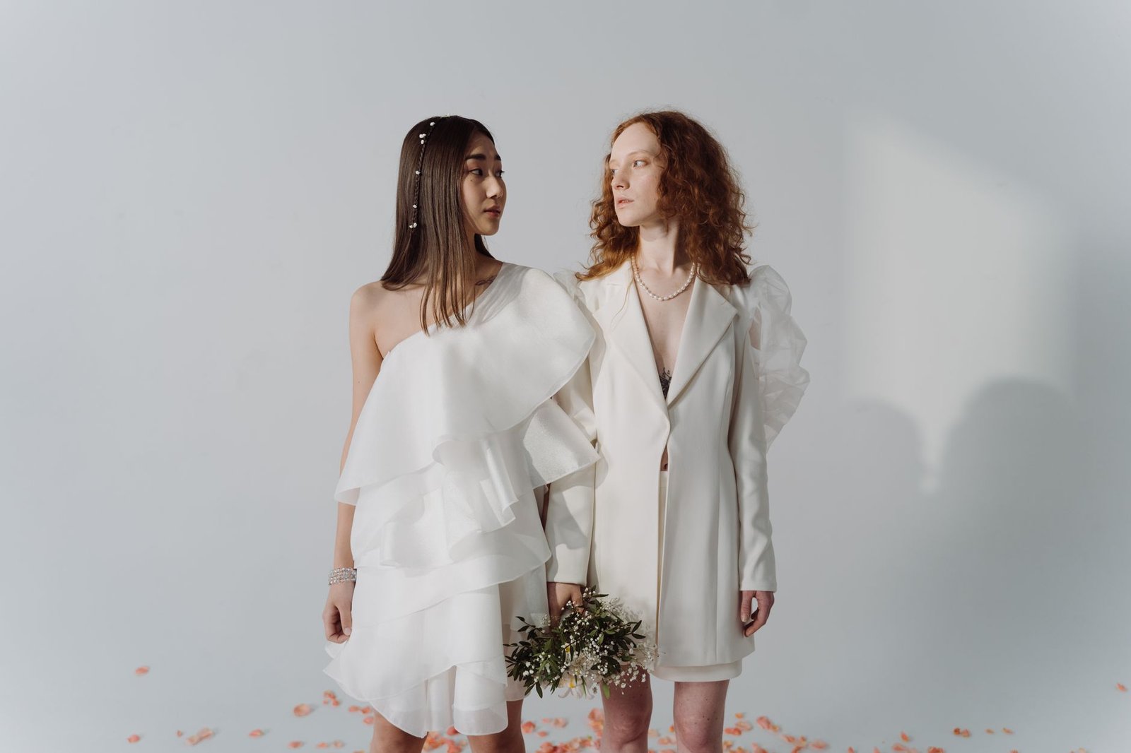 A stylish same-sex couple in white attire holding a bouquet during a studio wedding photoshoot.