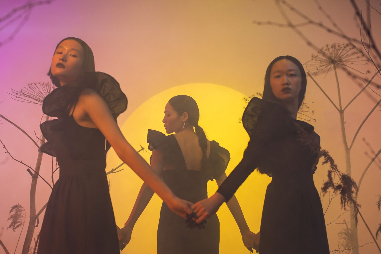 Artistic studio image of three Asian women holding hands, showcasing conceptual fashion with warm lighting.
