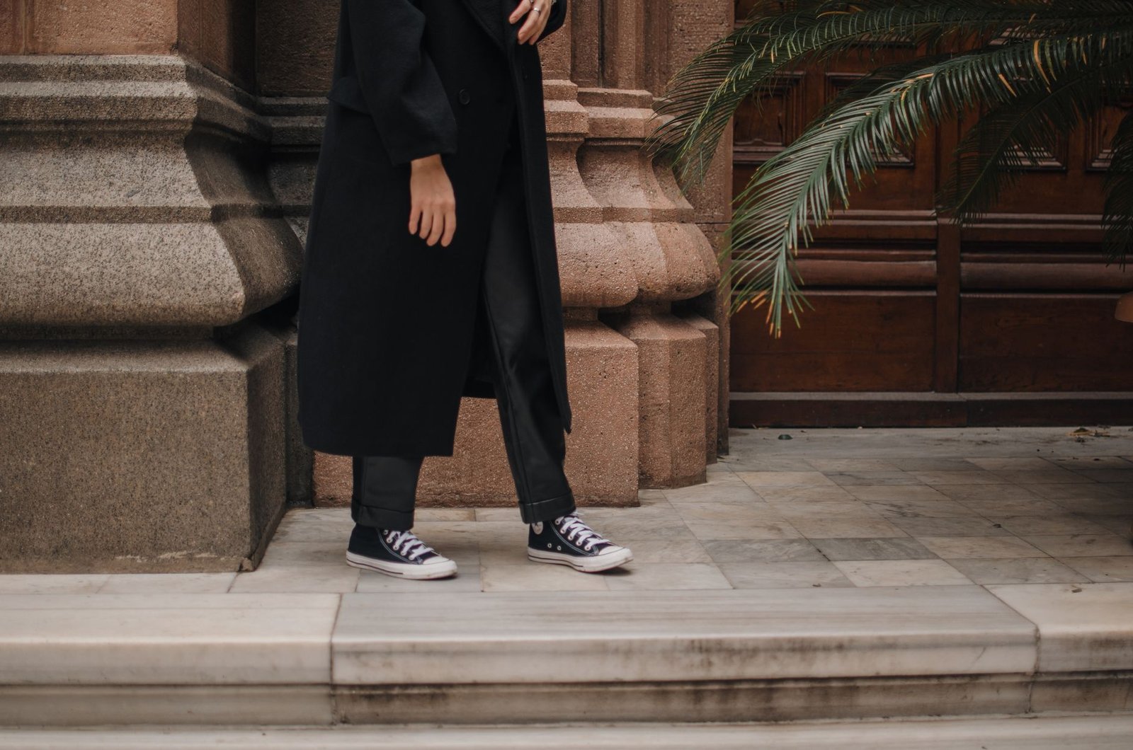 Crop view of unrecognizable person in black trousers and coat with sneakers standing on porch of stone building on daytime