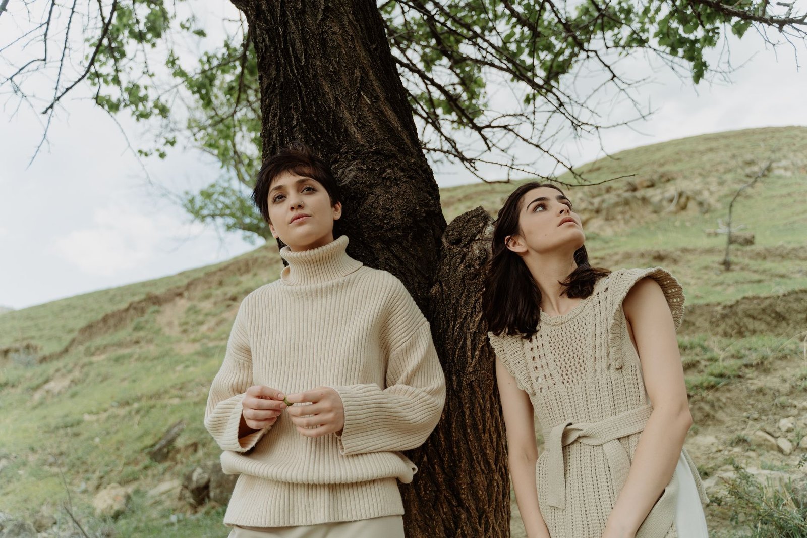Two women in stylish outfits leaning against a tree in a serene outdoor setting.