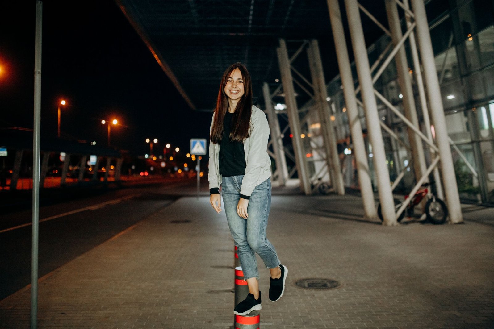 Full body cheerful female smiling and looking at camera while standing on fence post on street of city in evening