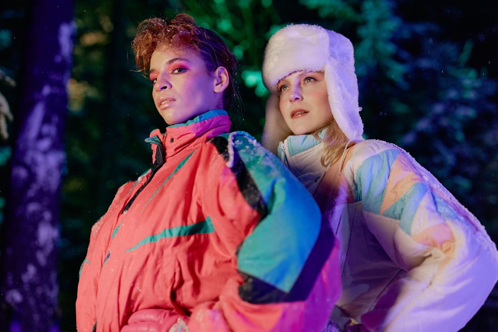 Two fashionable women posing in colorful winter jackets during a nighttime forest photoshoot.