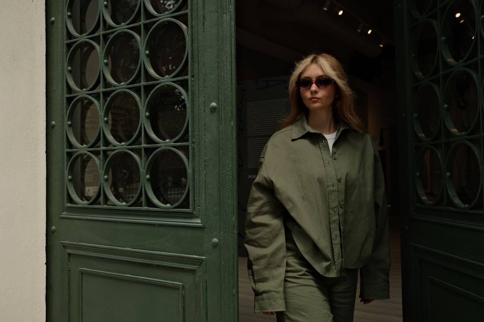 Woman in olive green oversized outfit and sunglasses exiting a vintage doorway.