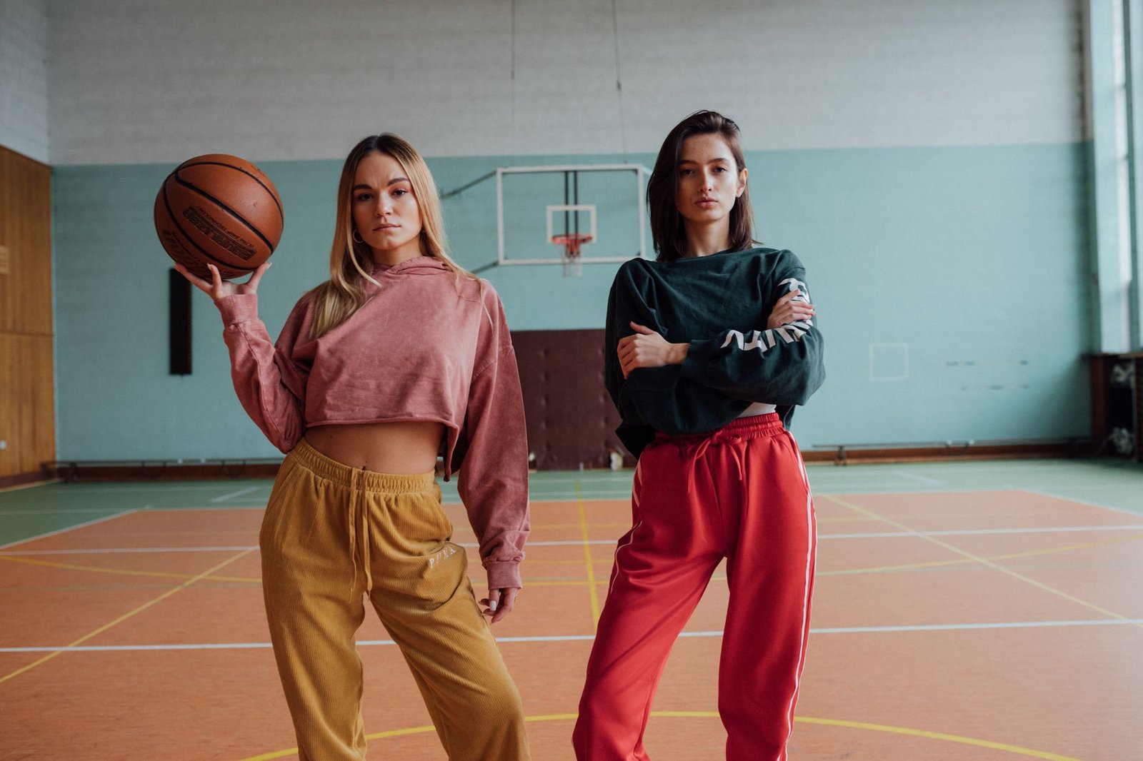 Two fashionable women pose on an indoor basketball court, embracing sporty style and attitude.