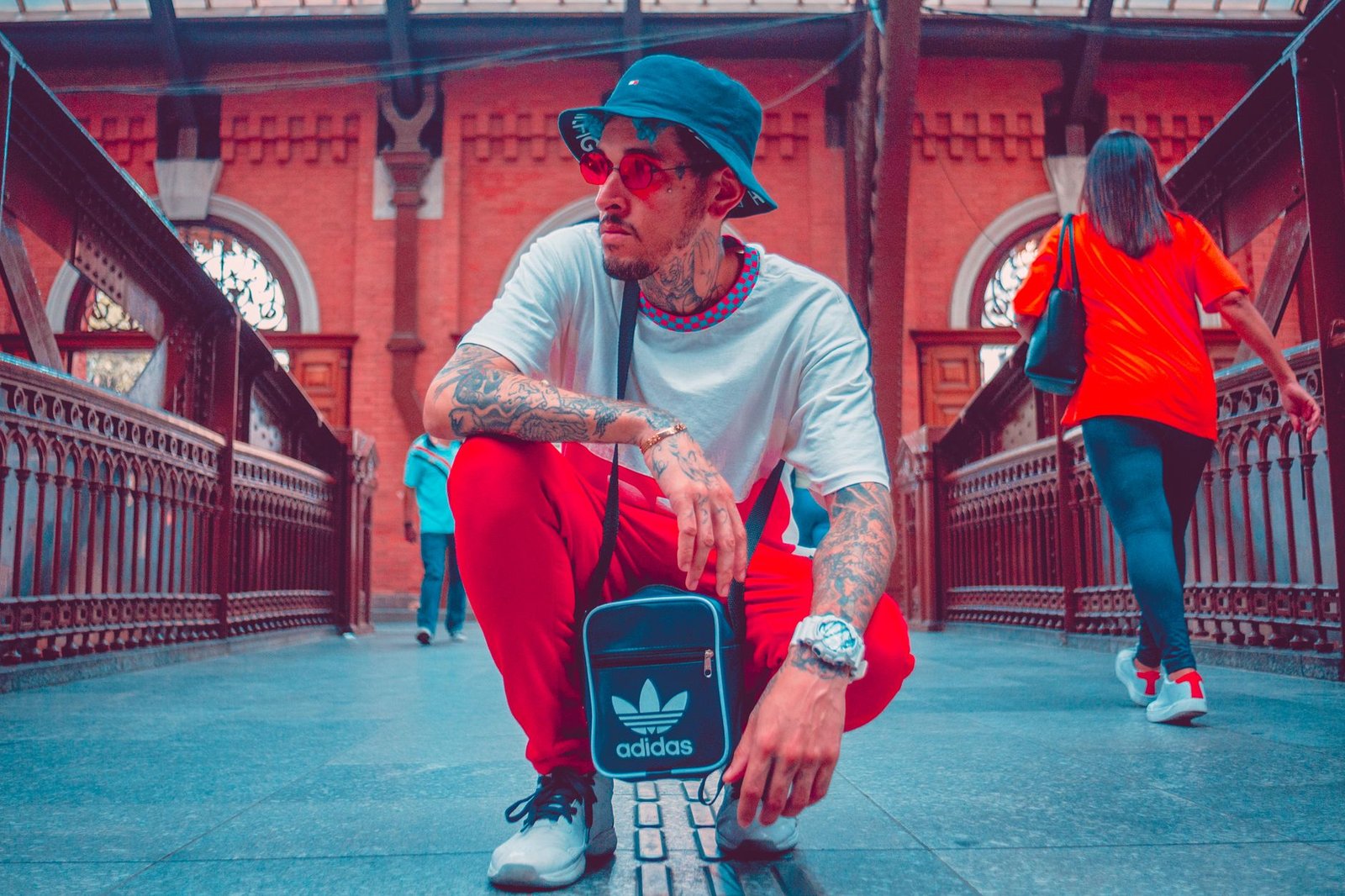 Fashionable tattooed man with sunglasses squatting on a bridge, showcasing urban style.