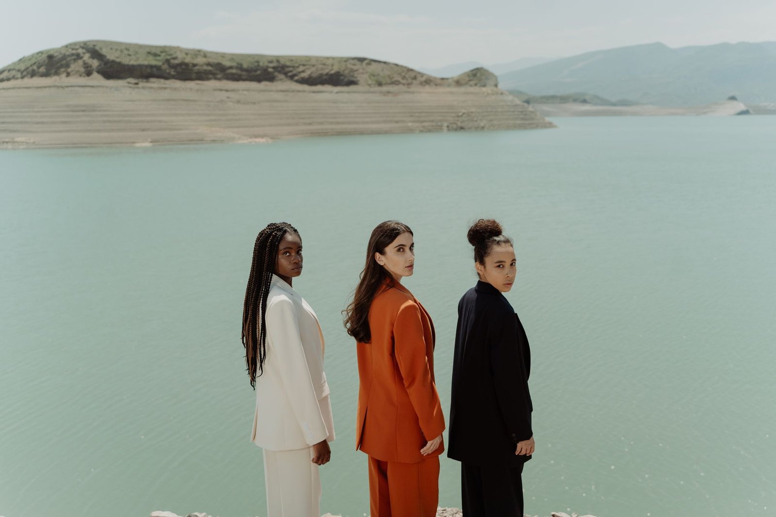 Three women in colorful blazers stand elegantly by a tranquil lake, exuding fashion and diversity.