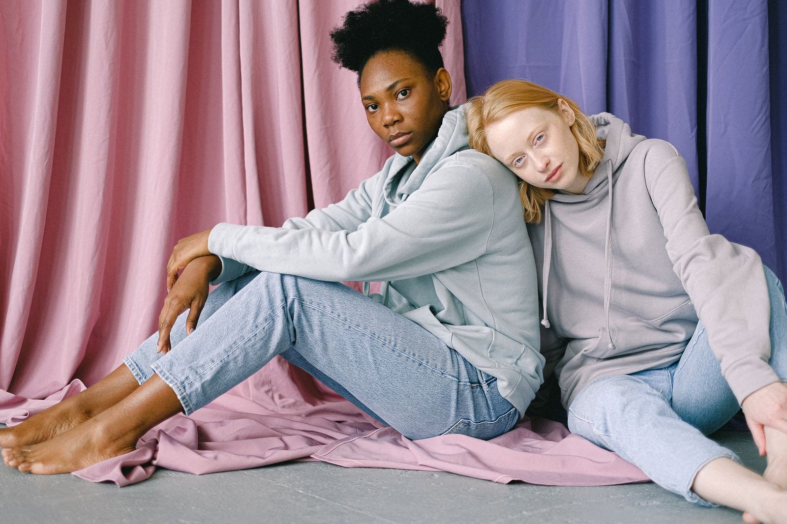 Two women sitting on the floor in jeans and hoodies, with a backdrop of pastel curtains.