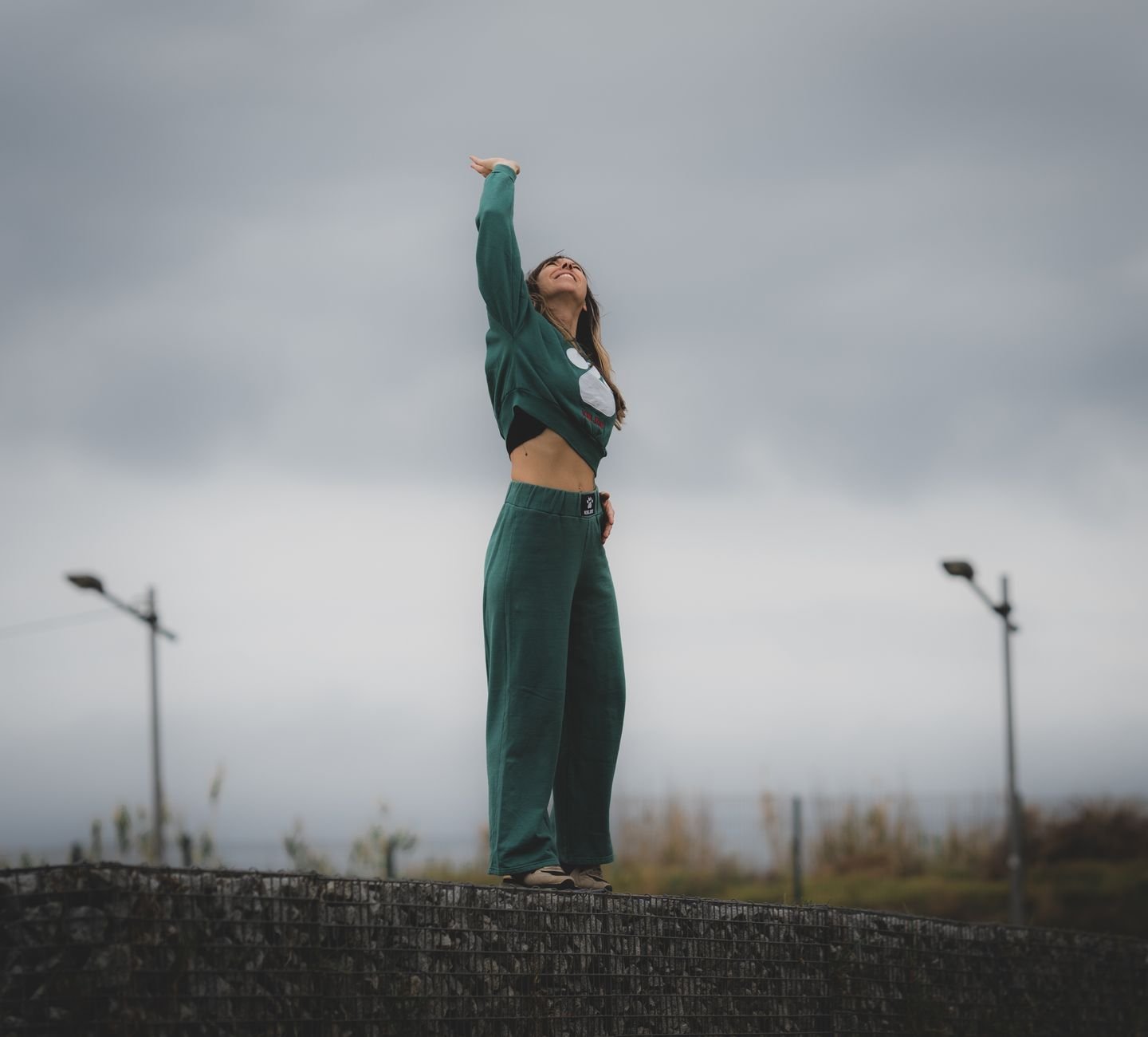 Young woman in green casual clothing standing outdoors with arm raised, set against a cloudy urban backdrop.