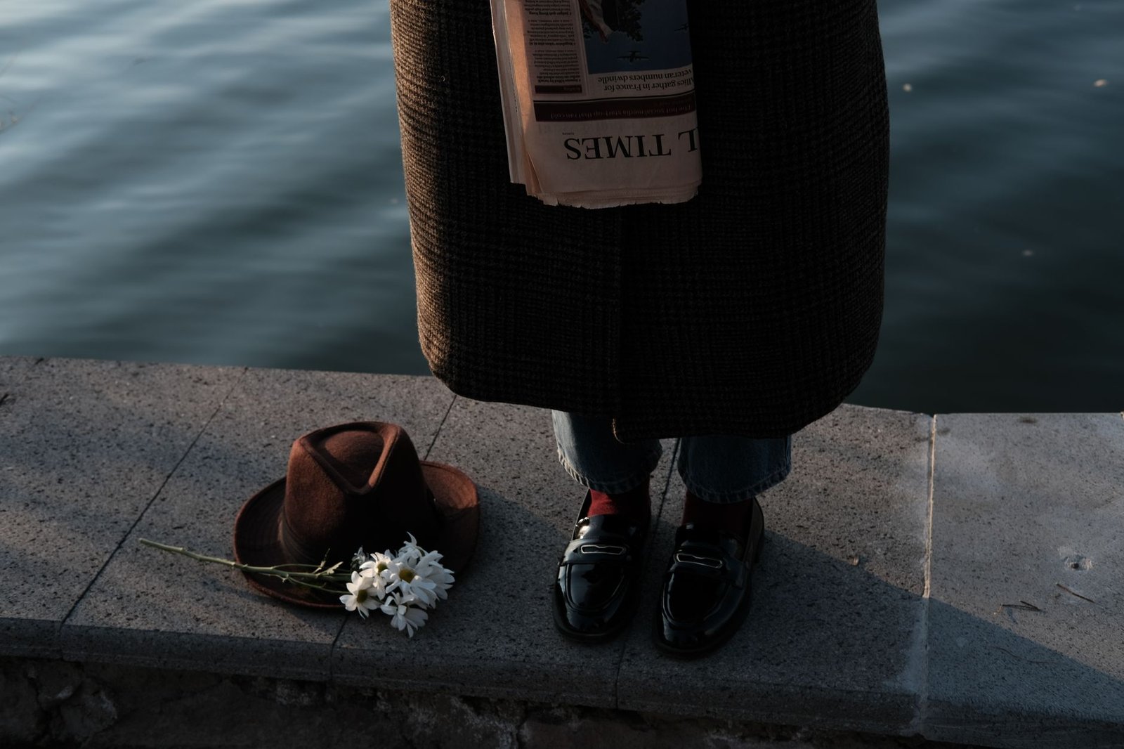A person stands by the water with a hat, flowers, and a newspaper, evoking a cinematic mood.