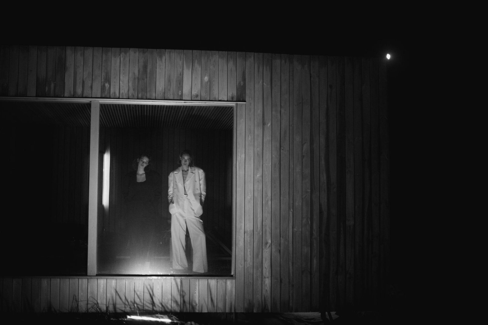 Black and white portrait of two individuals posing indoors, seen through a window at night.