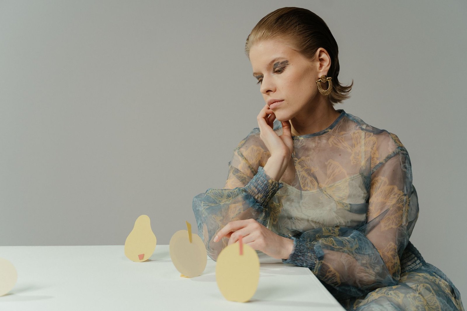 Stylish woman in sheer dress engaging with cut-out art on a table in a studio setting.