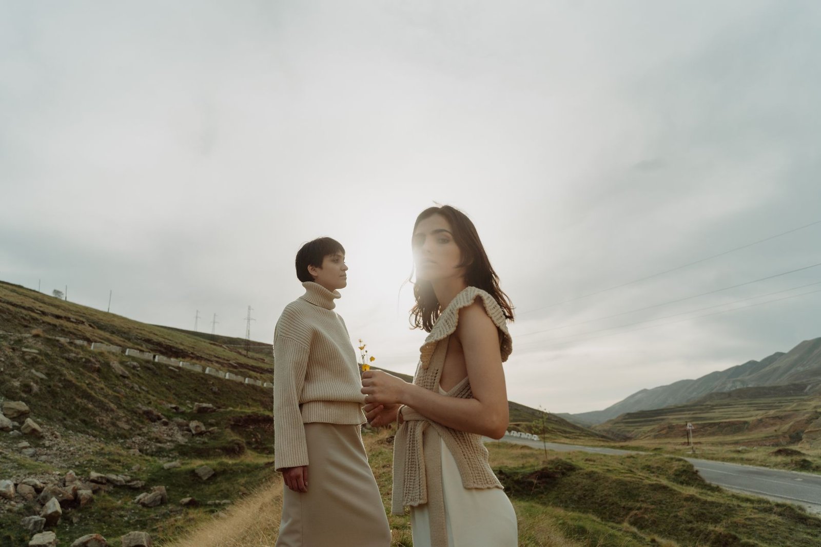 Two women pose in stylish outfits during a serene outdoor photoshoot with a mountainous backdrop.