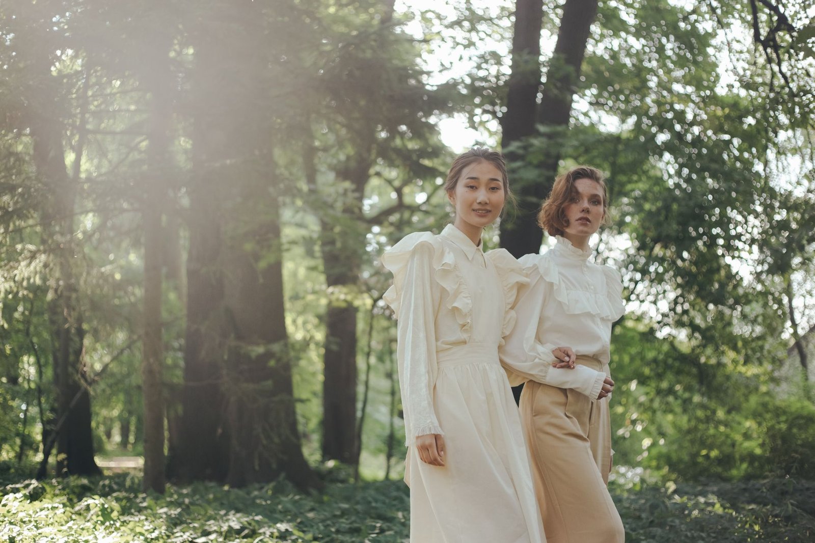 Two women in vintage attire walk gracefully through a serene forest setting.