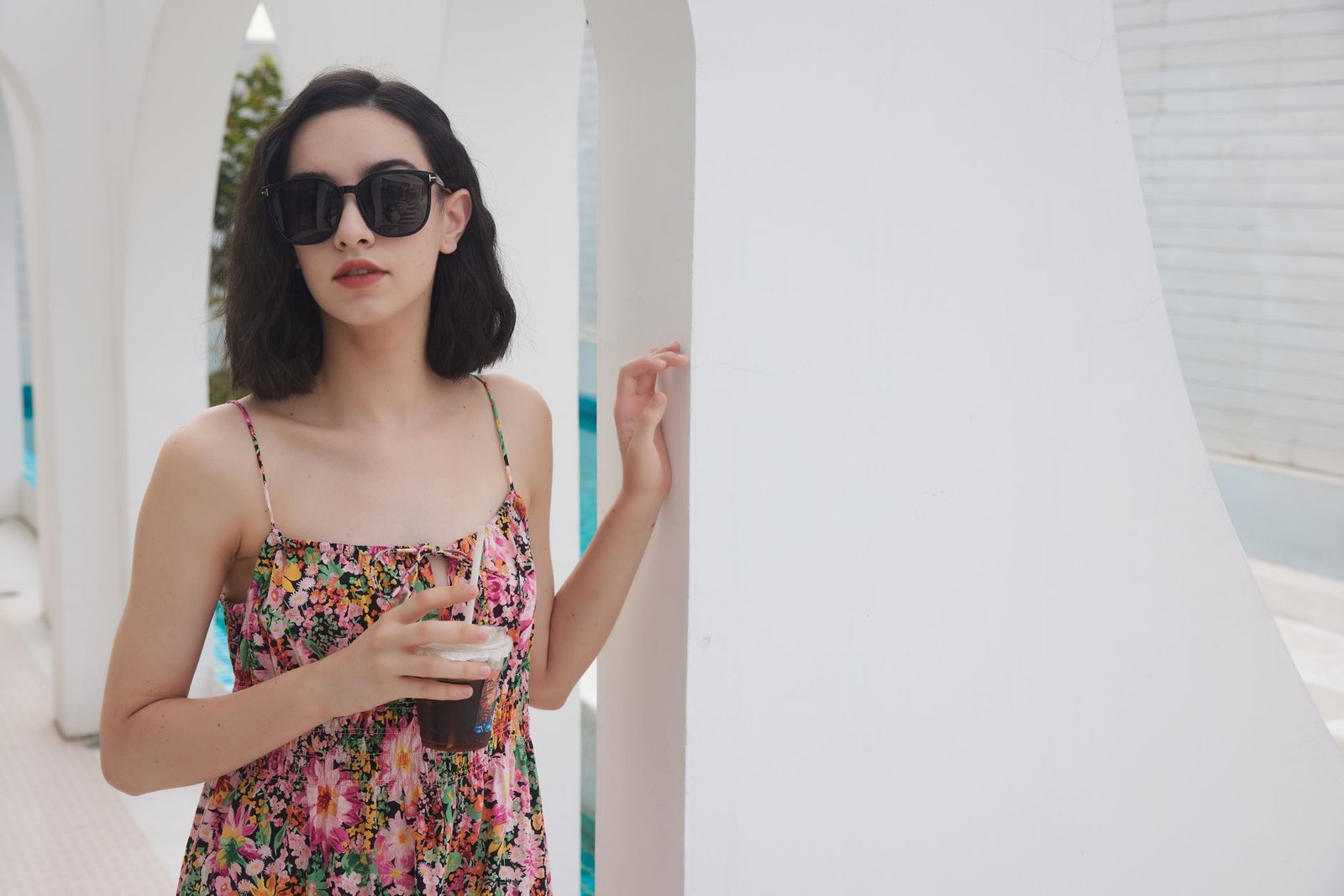 Elegant woman in floral summer dress and sunglasses holding a drink outdoors.