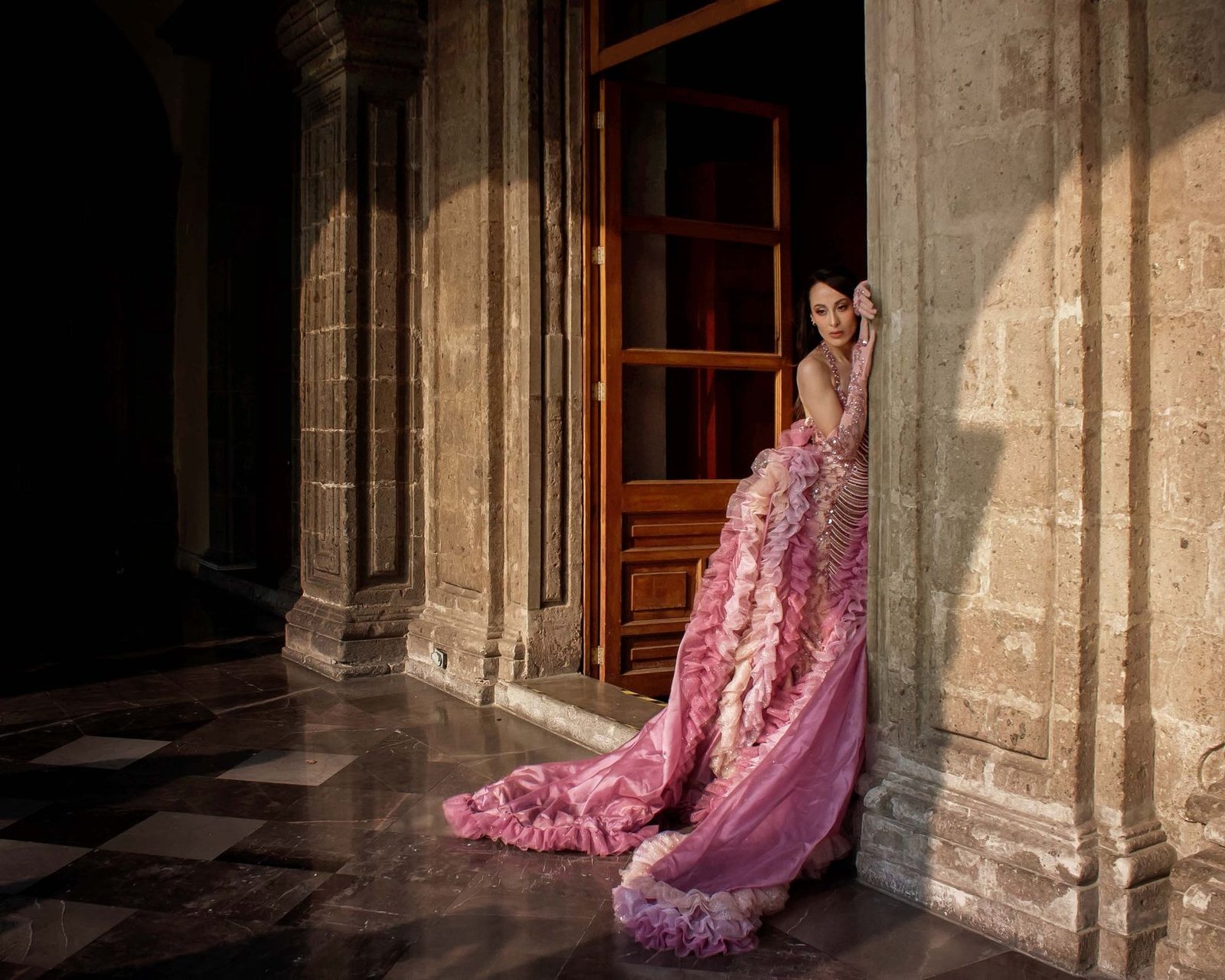 Elegant woman in pink ruffled gown leaning against ornate classical architecture.