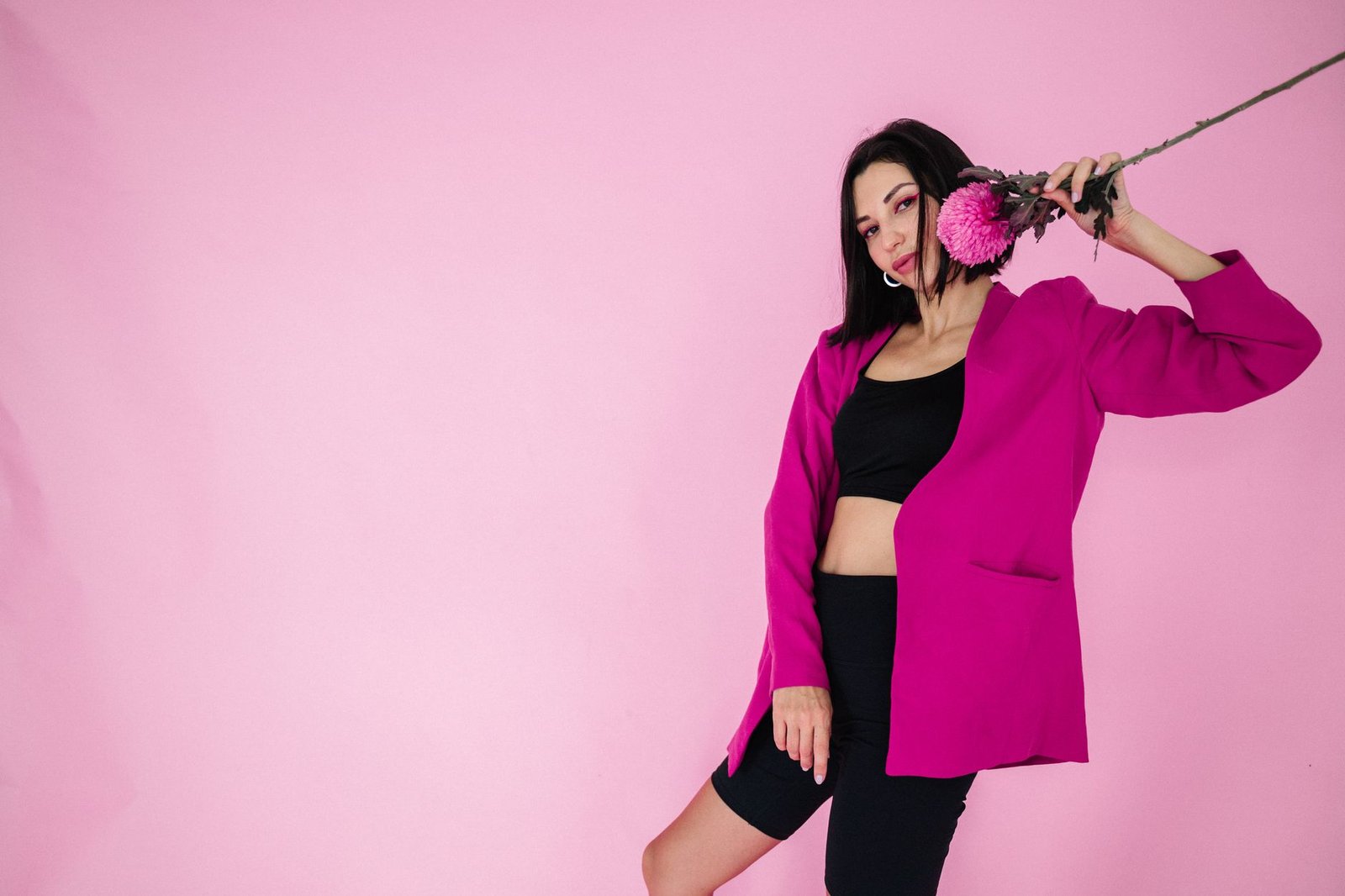 Fashionable woman in a bright fuchsia blazer holding a pink flower against a pink backdrop.