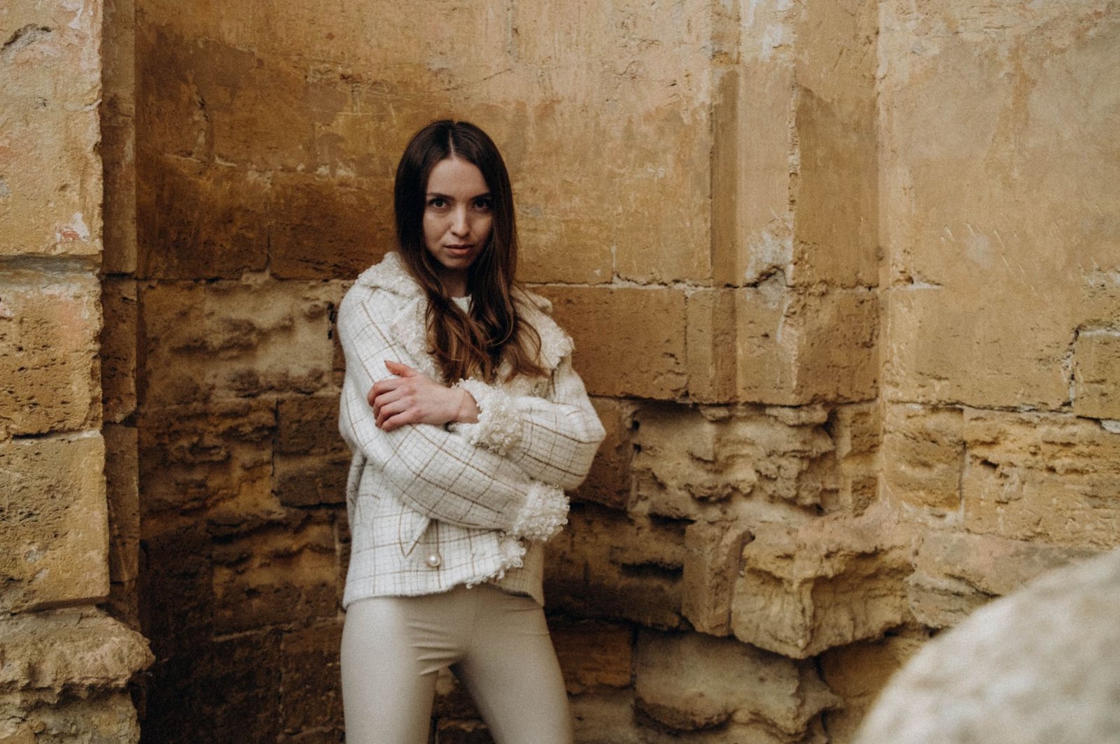 Fashionably dressed woman standing against a rustic stone wall, arms crossed confidently.