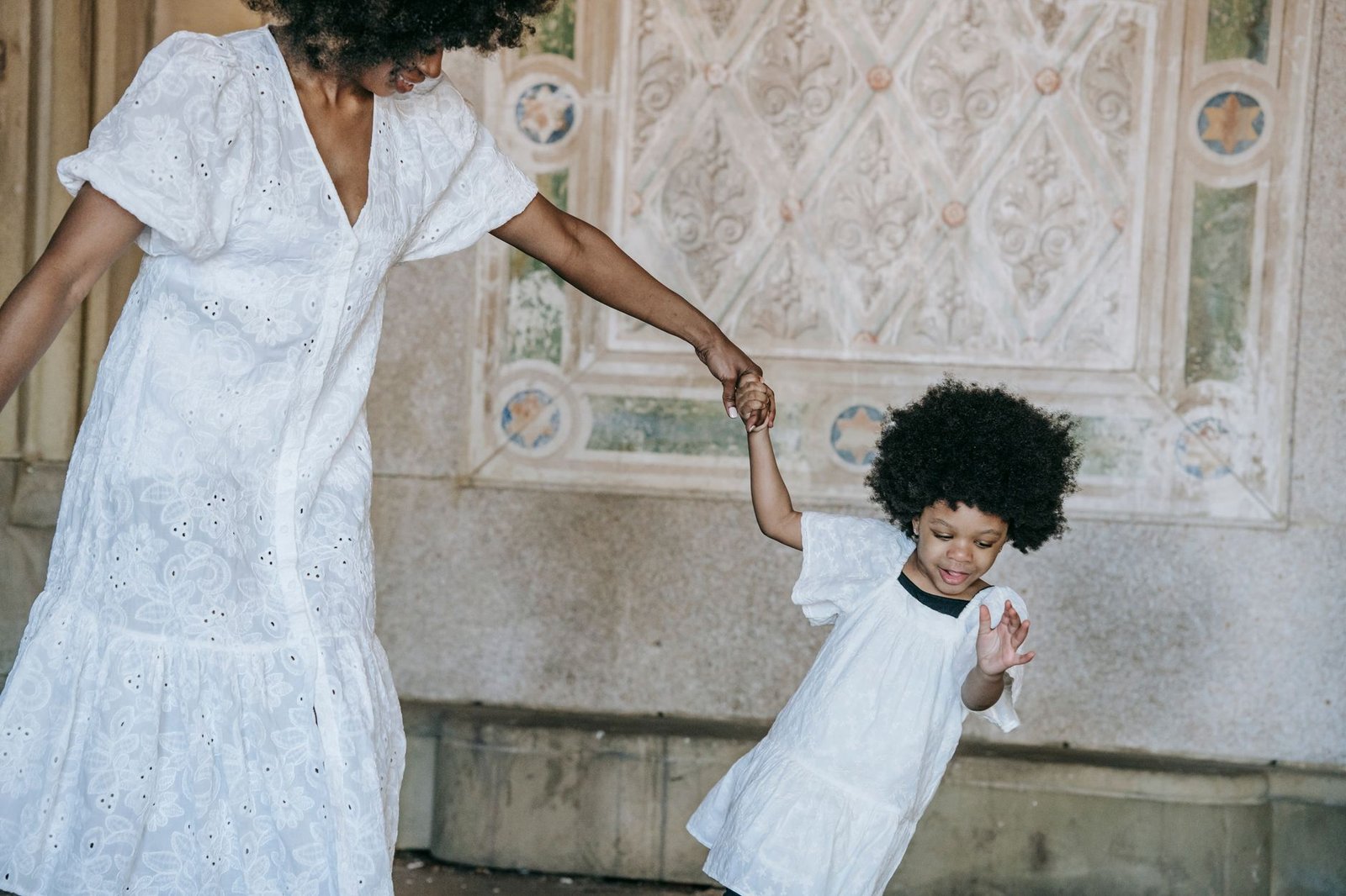 A happy mother and daughter holding hands and smiling while wearing white dresses indoors.