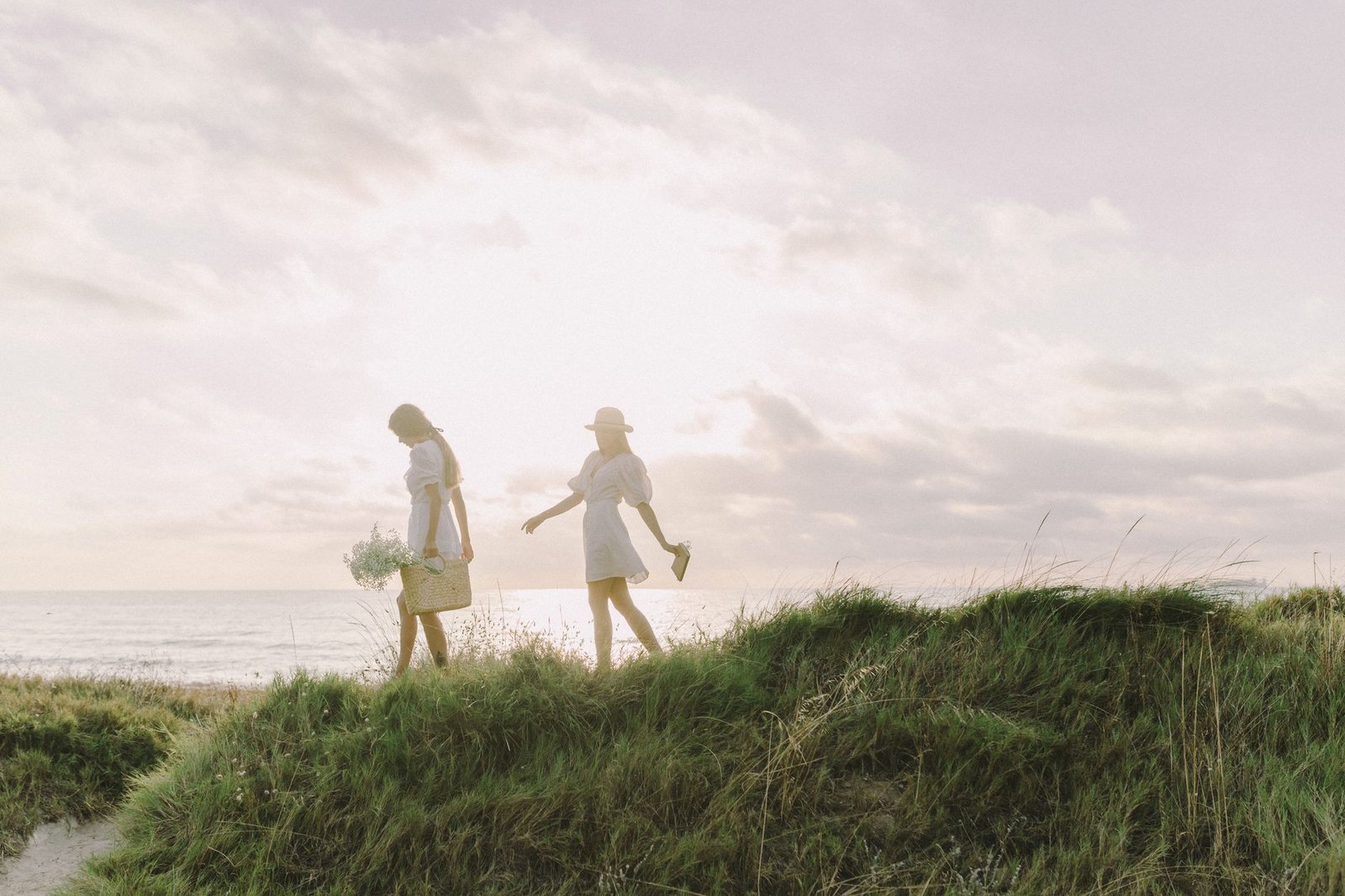 Two women walking on grassy dunes at the beach during a sunny summer day, capturing a bohemian vibe.