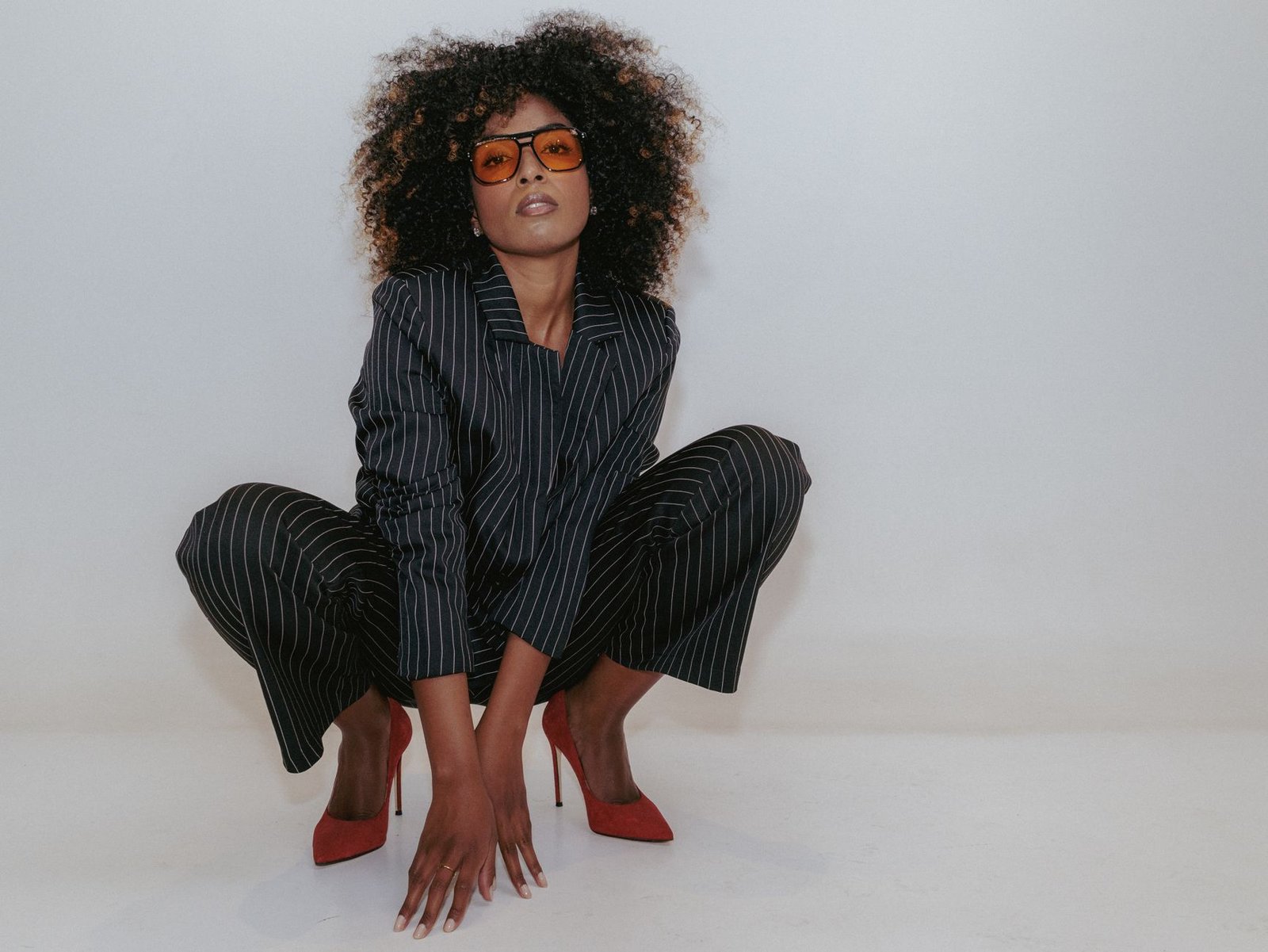 Confident woman in pinstripe suit and orange sunglasses posing indoors on white background.