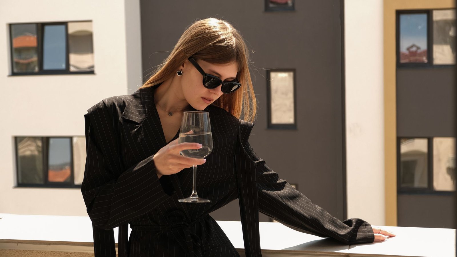 Stylish woman in sunglasses holding a drink outdoors with urban background.
