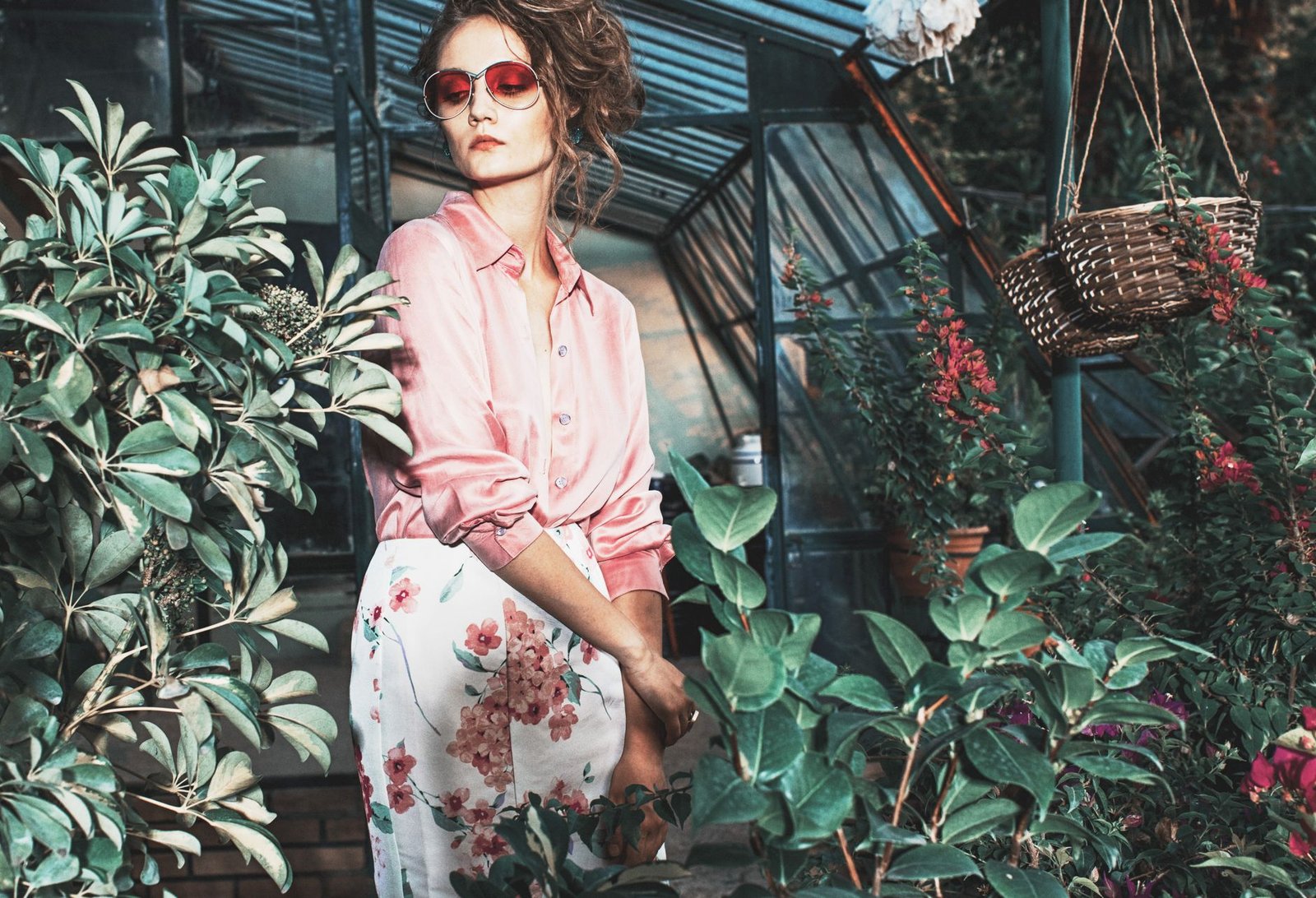 A fashionable woman in sunglasses and floral skirt poses elegantly in a greenhouse.