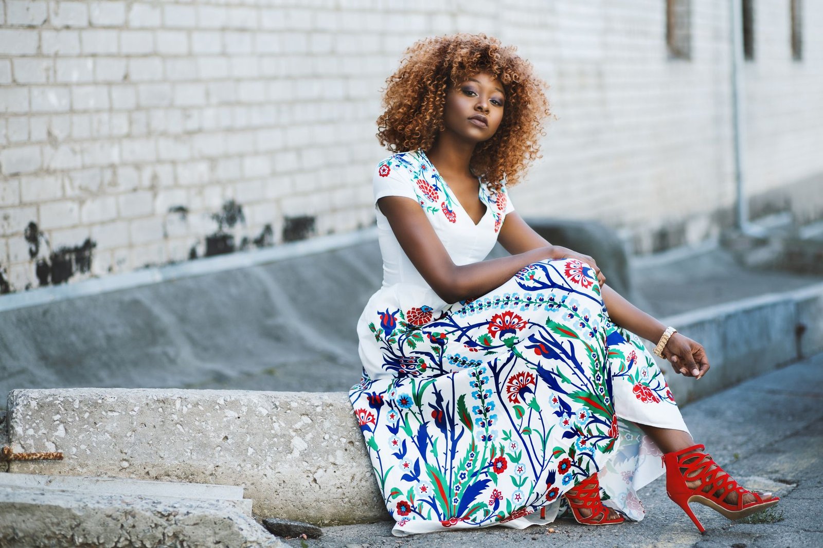 African American woman sitting and posing in a vibrant floral dress against a brick wall.