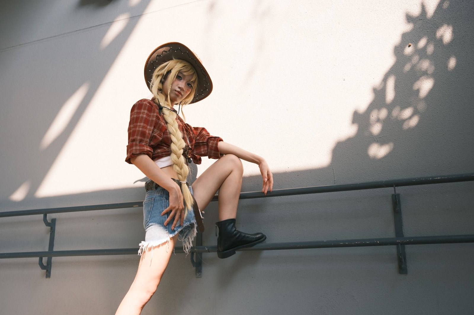 Stylish young woman posing in denim shorts and cowboy hat against a sunny wall.