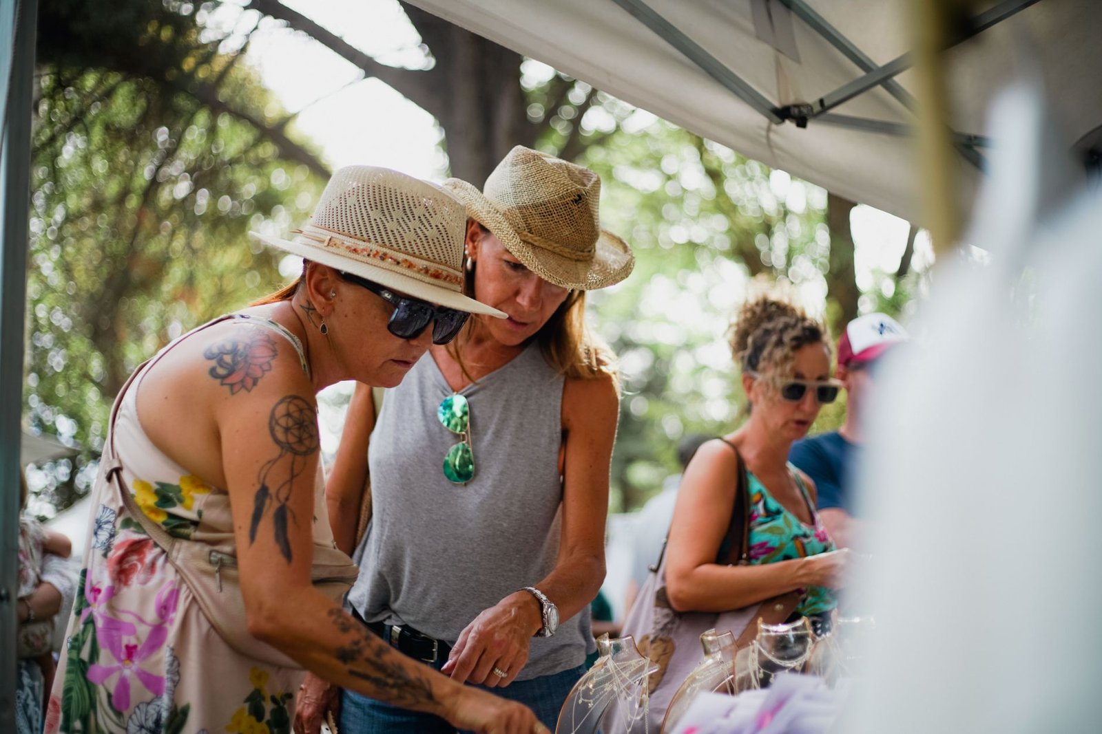Women browsing items at an outdoor market, wearing hats and sunglasses on a sunny day.